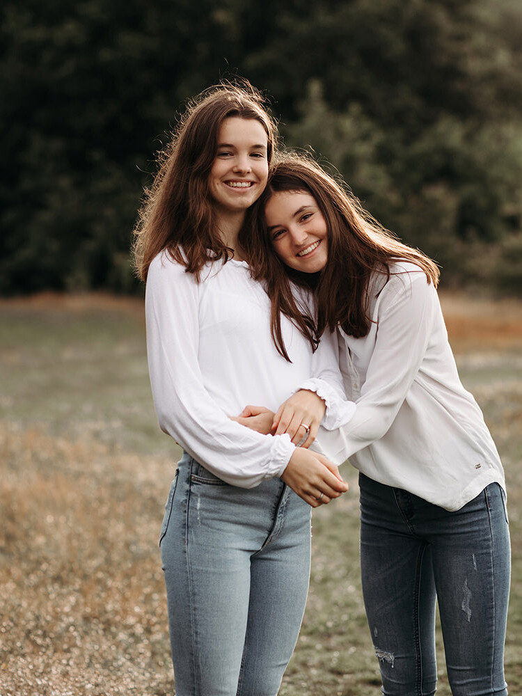 Twee lachende jonge zussen met lang bruin haar, witte tops en jeans knuffelen tijdens een zussen fotosessie buiten in de natuur in het Gooi, Blaricum