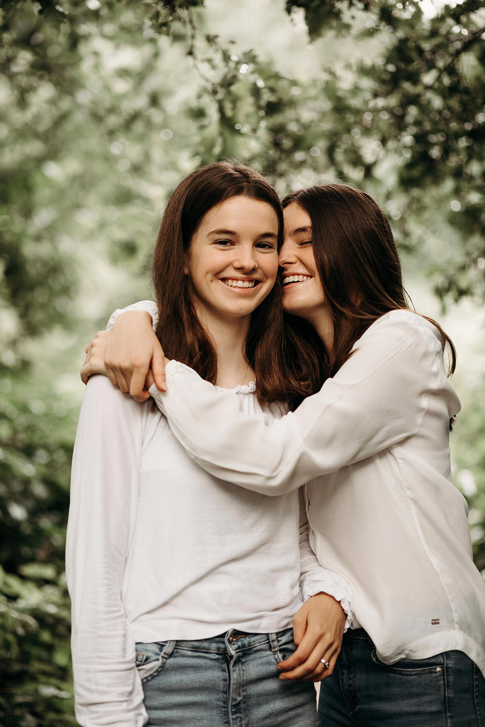 Twee bruinharige lachende jonge zussen poseren in een zussen portretfotosessie buiten in de Nederlandse natuur